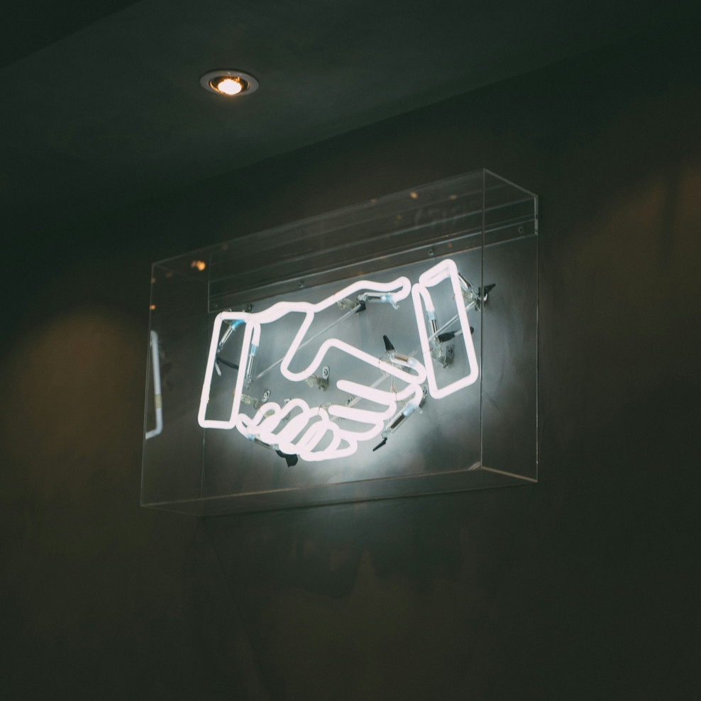 Leuchtendes Neon‑Symbol in Form zweier sich reichender Hände in einem transparenten Kasten an einer dunklen Wand. Darüber sind eingelassene Deckenstrahler zu sehen, die den Raum dezent beleuchten.