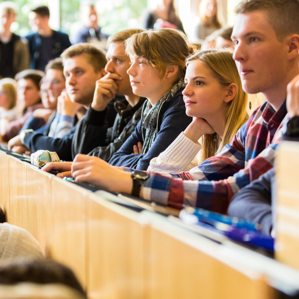 Auf dem Foto sitzen Studierende in einem vollen Hörsaal und hören aufmerksam zu. Wer zu in ihnen spricht, ist nicht zu erkennen.
