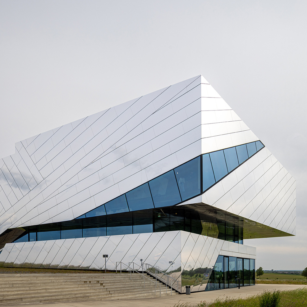 Futuristisches, silbrig glänzendes Gebäude mit schrägen, kantigen Fassaden und langen Glasfronten. Das paläon steht frei in der Landschaft, umgeben von weitem Himmel und Wiesen.