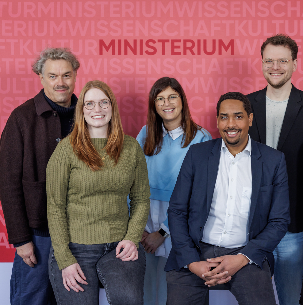Sieben Personen posieren vor einer roten Wand mit dem Schriftzug ‚Ministerium‘. Zwei Personen sitzen vorn, fünf stehen dahinter. Die Gruppe blickt in die Kamera, eine Person hält mehrere Bücher im Arm.