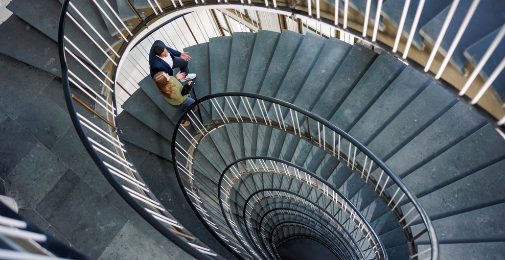Blick von oben in ein helles Treppenhaus mit einer geschwungenen Wendeltreppe. Zwei Personen stehen auf einer Treppenstufe und unterhalten sich. Das spiralförmige Geländer und die Treppenläufe prägen die Aufnahme.