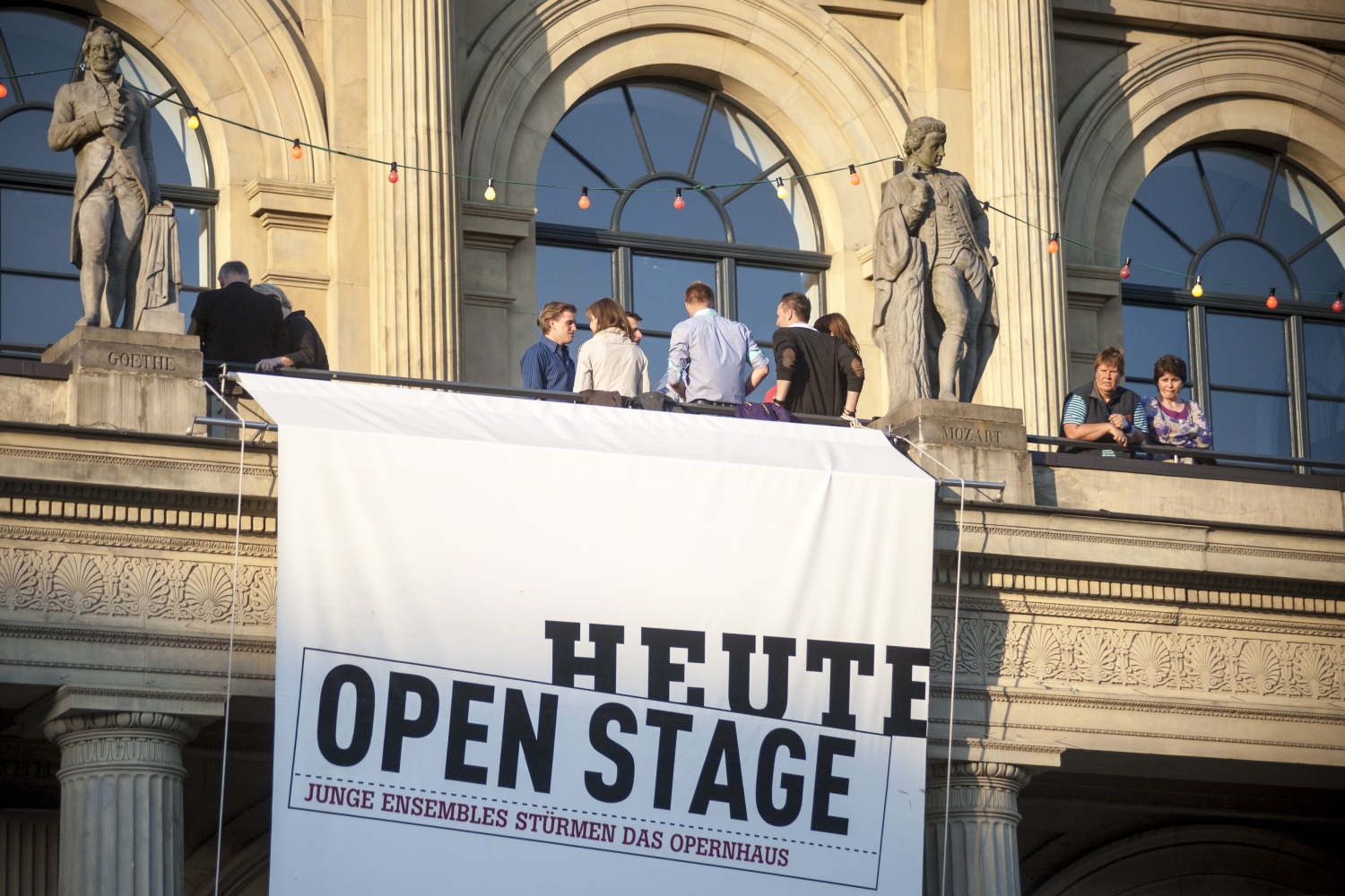 Auf dem Balkon des Opernhauses in Hannover stehen Menschen. An der Ballustrade ist ein Banner angebracht, auf dem "Heute Open Stage. Junge Ensembles stürmen das Opernhaus" steht.