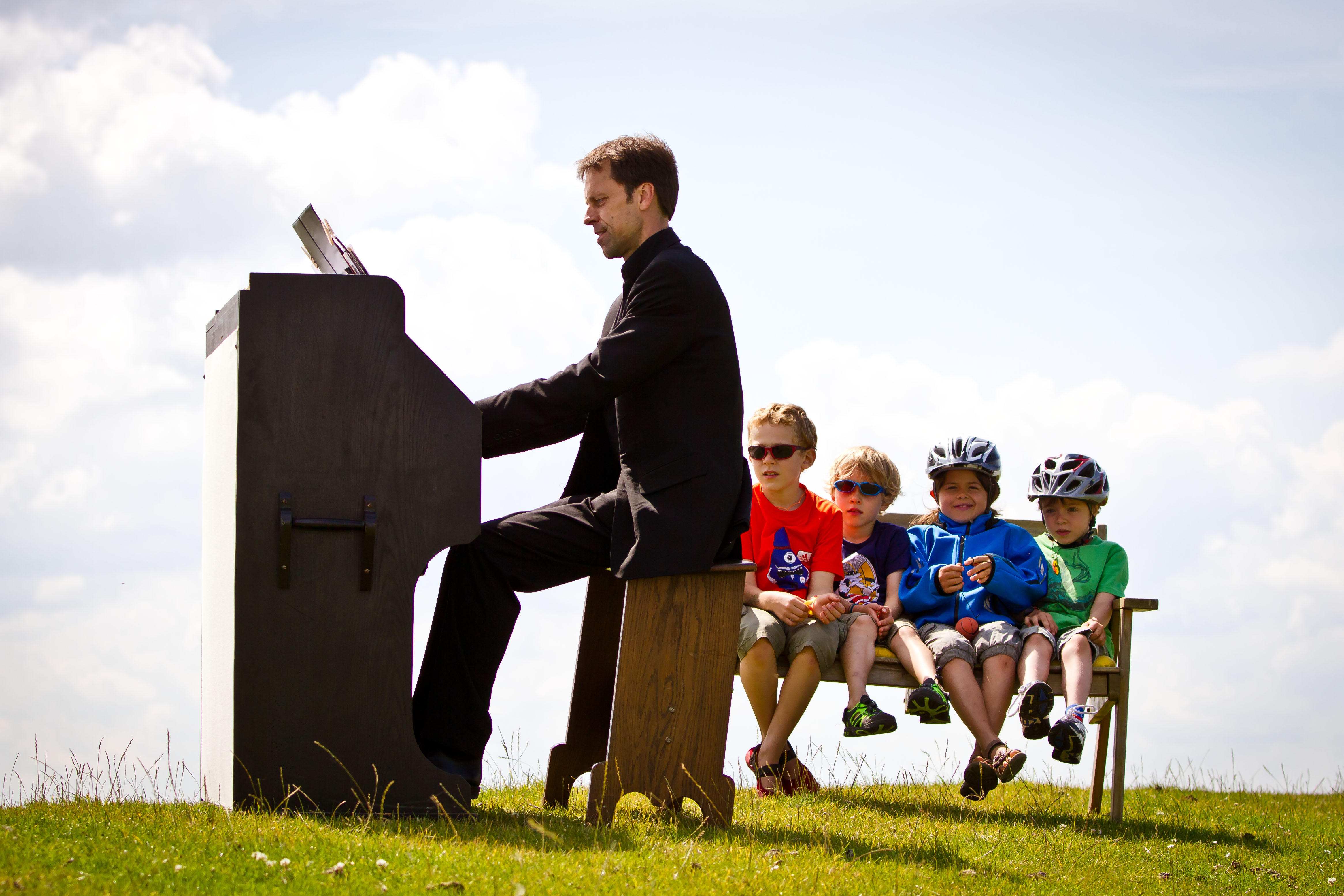 Auf einer Wiese sitzt ein schwarz gekleideter Pianist am Klavier. Hinter ihm vier Kinder in bunter Kleidung auf einer Bank, zwei von ihnen tragen einen Fahrradhelm.